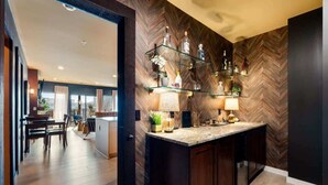 A modern kitchen with a herringbone wood wall, a glass shelf with bottles, and a dining area with a table and chairs.