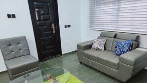 A living room with a grey sofa, a grey armchair, and a glass coffee table with a tree stump centerpiece. - Super Deluxe Room #1 (Ikeja)