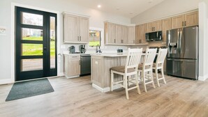 A modern kitchen with light wood cabinets, a stainless steel refrigerator, and a breakfast bar with chairs. - Sunset Sky Lodge (McHenry)