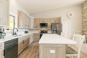 A modern kitchen with wooden cabinets, stainless steel appliances, a large island, and a dining area with white chairs. - Sunset Sky Lodge (McHenry)
