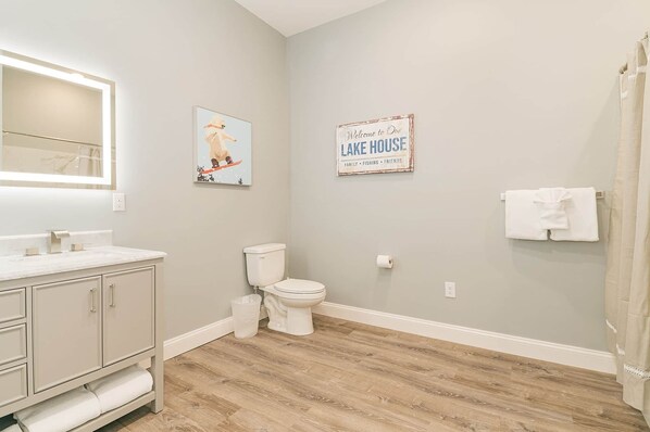 A bathroom with a toilet, sink, mirror, and a 'Welcome to Our Lake House' sign.
