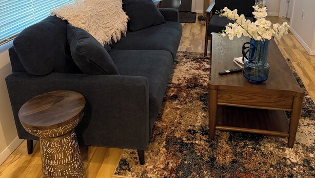 A living room with a dark grey sofa, a wooden coffee table with a vase of white flowers, a patterned rug, and a ceiling fan.
