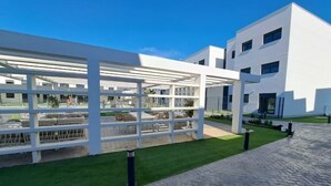 A modern building with a white exterior, a covered outdoor area with seating, and a paved walkway. - Newbuild apartment Estepona pool - beach - gym (Estepona)