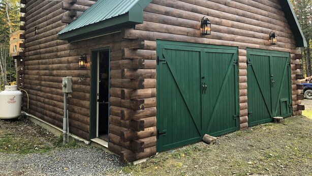 Una cabaña de troncos con techo de metal verde, dos puertas de garaje verdes y un pequeño porche.