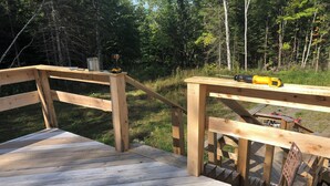 Una terraza de madera con una silla metálica y una mesa, rodeada de un bosque.