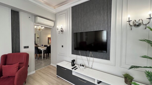 Ein modernes Wohnzimmer mit einem großen Flachbildfernseher an einer Wand mit vertikaler Wandverkleidung, einem weißen Konsolentisch mit TV-Standfuß, einem roten Sessel und einem Essbereich im Hintergrund.