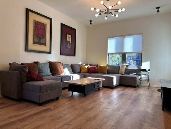 A modern living room with a sectional sofa, a coffee table, and a bar stool. There are two framed artworks on the wall and a large window providing natural light.
