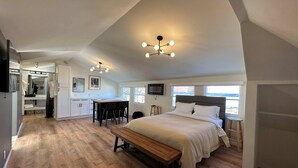 Un dormitorio amplio con una cama grande, un banco de madera, una mesa de comedor con sillas, una zona de cocina y un televisor montado en la pared.