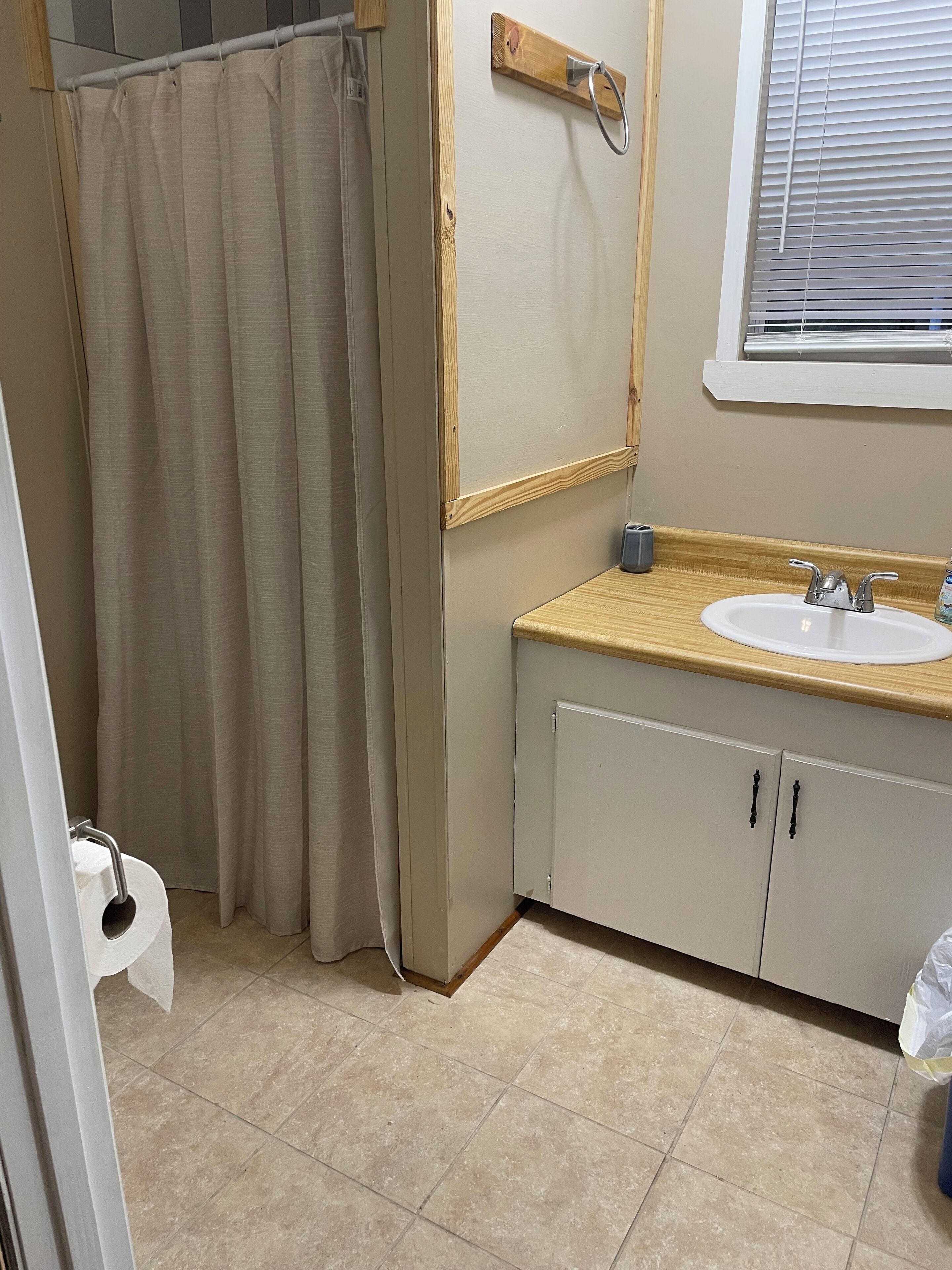 Une salle de bain avec un rideau de douche beige, un miroir encadré en bois, un lavabo avec un plan de toilette en bois et une fenêtre avec des stores.