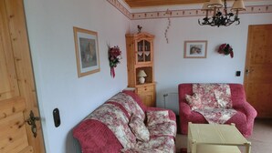 Ein gemütliches Wohnzimmer mit Holzboden, einer Couch, einem Sessel, einem kleinen Tisch und einem Schrank.