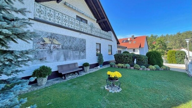 Ein zweistöckiges Haus mit geneigtem Dach, einem Balkon mit dekorativen Geländern und einem gepflegten Rasen mit einer Bank und Blumentöpfen.
