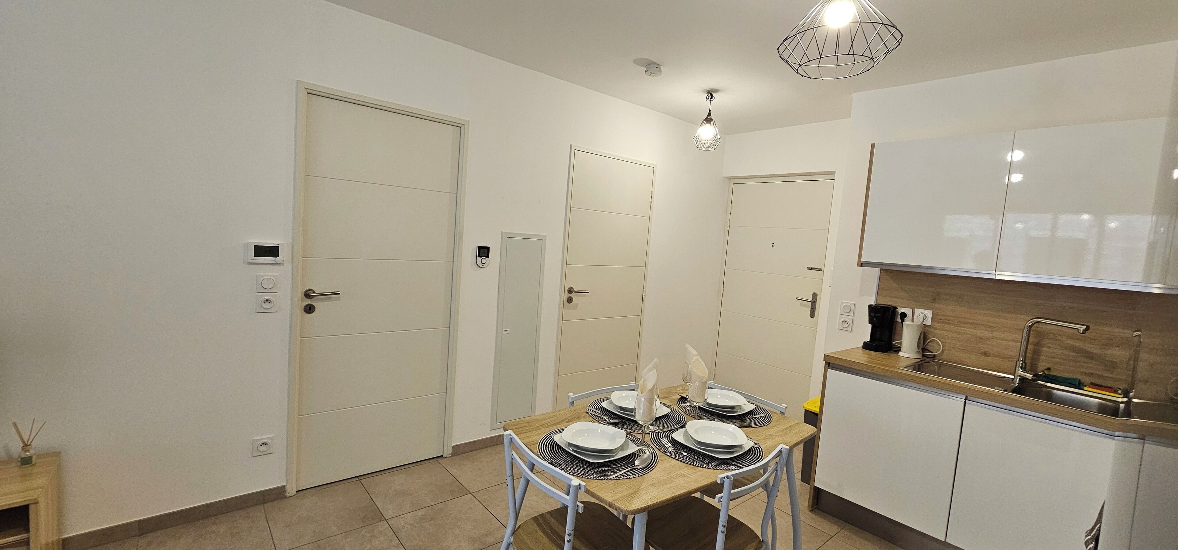 Une cuisine compacte avec des meubles blancs, une table à manger en bois pour quatre personnes et un luminaire suspendu moderne.