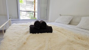 A bedroom with a bed covered in a fluffy white blanket, a window with a view of buildings, and a radiator on the wall. - Cosy Apartment In A Green Setting -Nice (Nice)