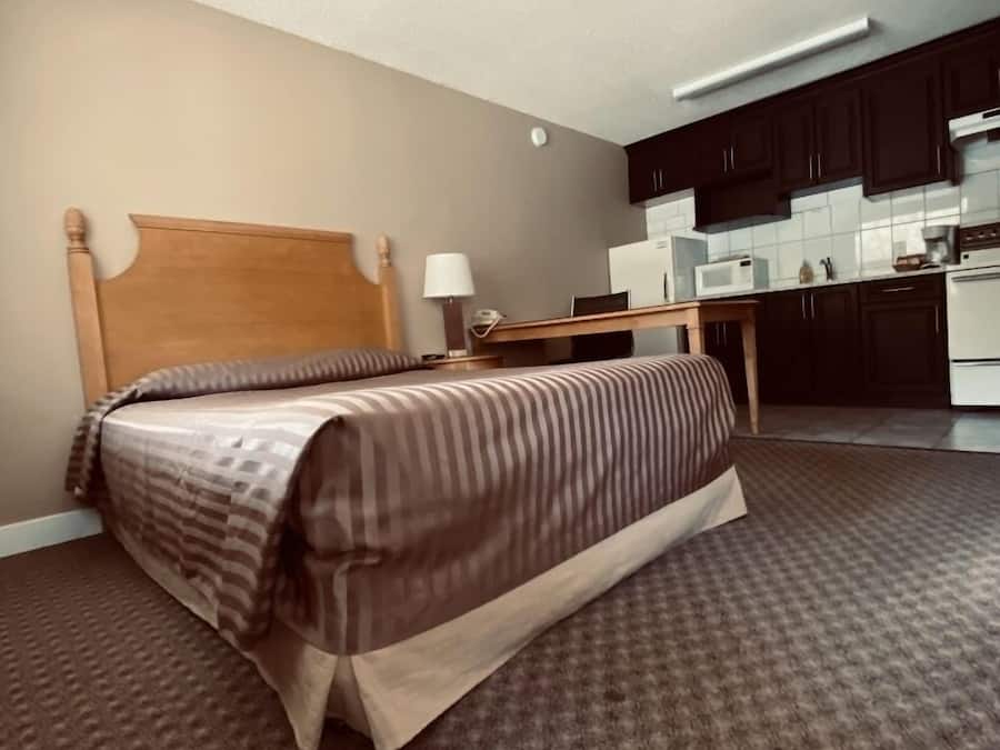 Un dormitorio con cama, mesita de noche, lámpara y una kitchenette al fondo.