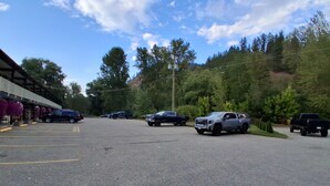 A parking lot with several vehicles, a building with a covered area on the left, and a forested hillside in the background. - Twin Creeks Motel (Lumby)