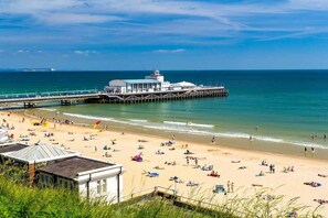 A beach with a pier extending into the ocean, a small building near the shore, and people relaxing on the sand.