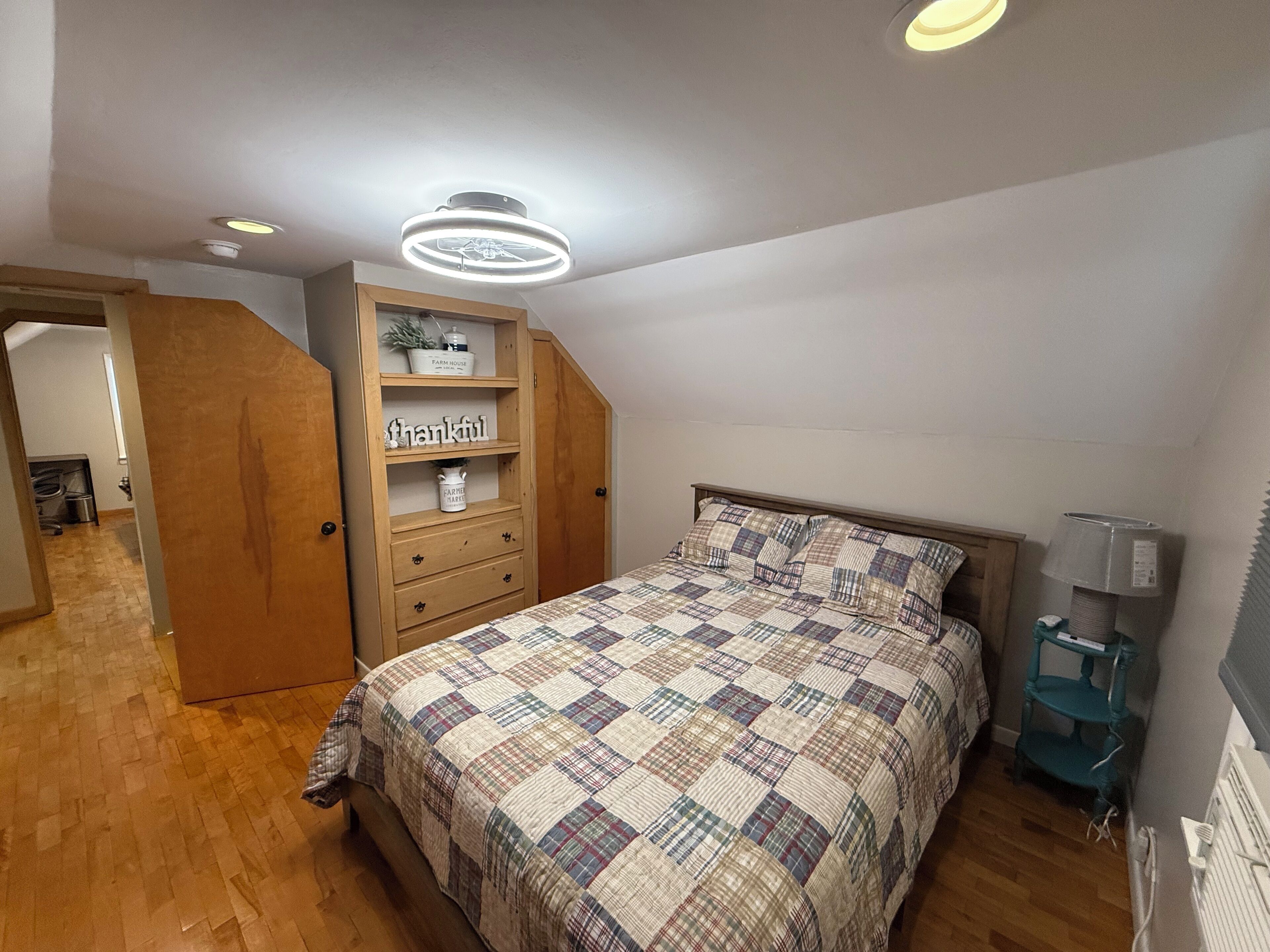 A bedroom with a wooden wardrobe, a bed with a plaid quilt, and a ceiling light fixture.