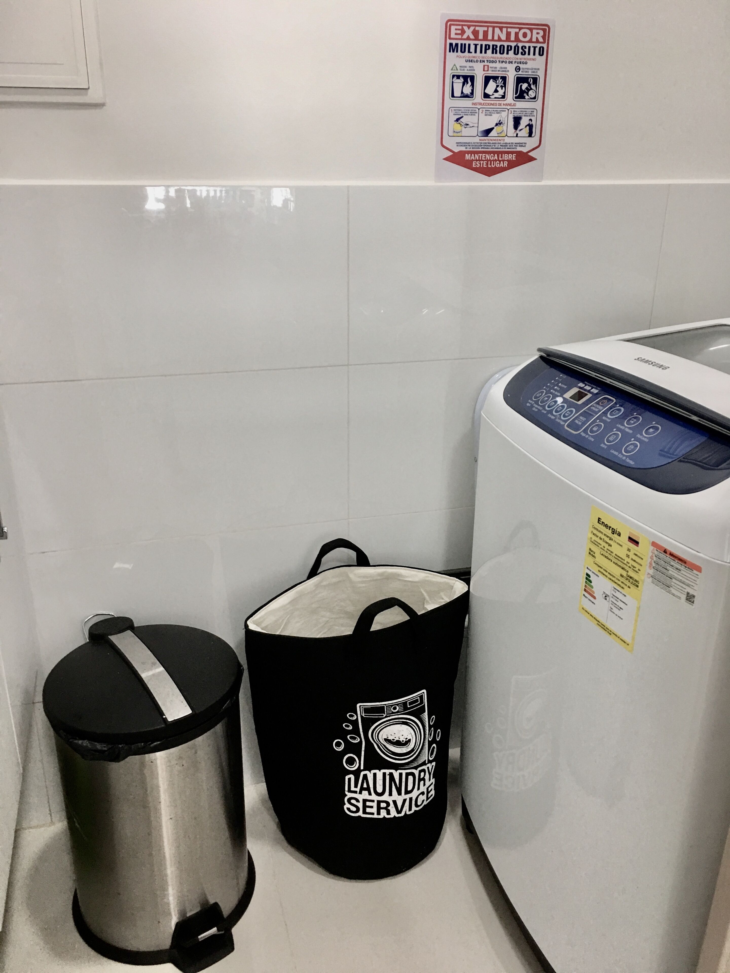 A laundry service bag, a trash can, and a washing machine in a room with white tiled walls.