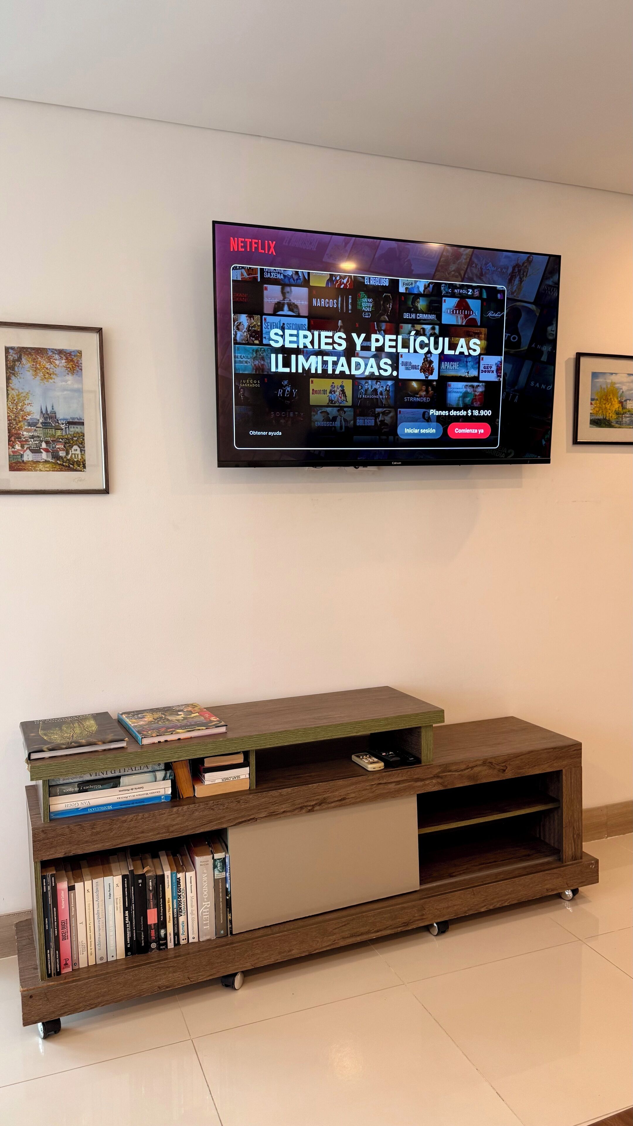 A flat-screen TV mounted on a wall displaying a Netflix interface with various series and movies. Below the TV, there is a wooden entertainment unit with books and a remote control.