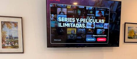 A flat-screen TV mounted on a wall displaying a Netflix interface with various series and movies. Below the TV, there is a wooden entertainment unit with books and a remote control.