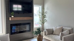A modern living room with a flat-screen TV, a fireplace, a ceiling fan, and a large window.