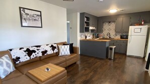 A modern living room with a brown leather sofa, a wooden coffee table, and a patterned throw pillow. A kitchen with grey cabinets and a white refrigerator is visible in the background. - Minutes to the Square • Fire Pit • Dog Friendly (Mountain View)