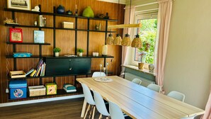 Un coin repas avec une table en bois, des chaises blanches, un meuble noir avec des étagères et une fenêtre avec des rideaux.