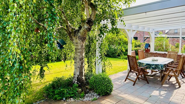 Une terrasse avec une table ronde et des chaises, entourée de verdure et surmontée d’une pergola blanche.