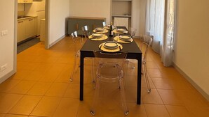 Een moderne eetruimte met een zwarte tafel voor vier personen, transparante stoelen, een ronde plafondlamp en een keuken zichtbaar door een open deur.
