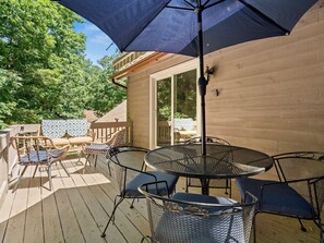 Eine Holzterrasse mit einem runden Metalltisch und Stühlen, einem großen Sonnenschirm und Blick auf ein Haus und Bäume.