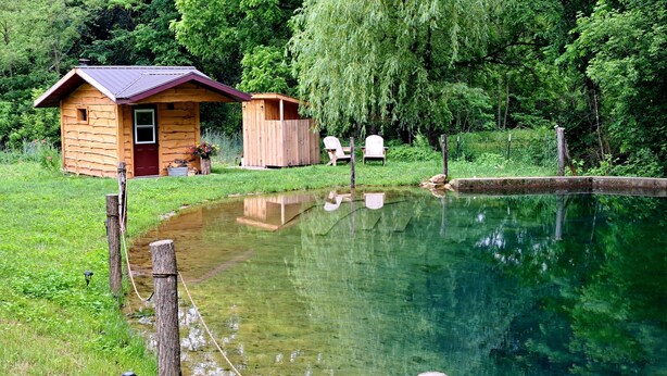 Een houten hutje met een klein vijvertje, omringd door weelderig groen en een houten terras met stoelen.