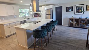 A modern kitchen with a central island, bar stools, and a dining area with a cabinet displaying items. - Charming 2-bedroom house in New Port Richey with WiFi, AC. Close to downtown (New Port Richey)