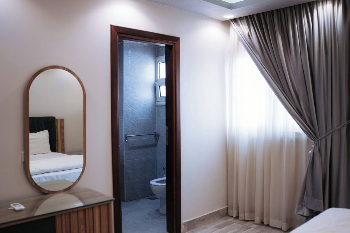 Habitación de hotel con mesita de noche de madera, un espejo, cama, baño con inodoro y ducha, y ventana con cortinas.