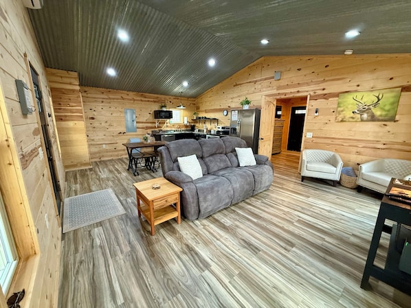 Interior de uma cabana de toras com um sofá cinza, mesa de jantar de madeira, área de cozinha e um quadro de um veado na parede.