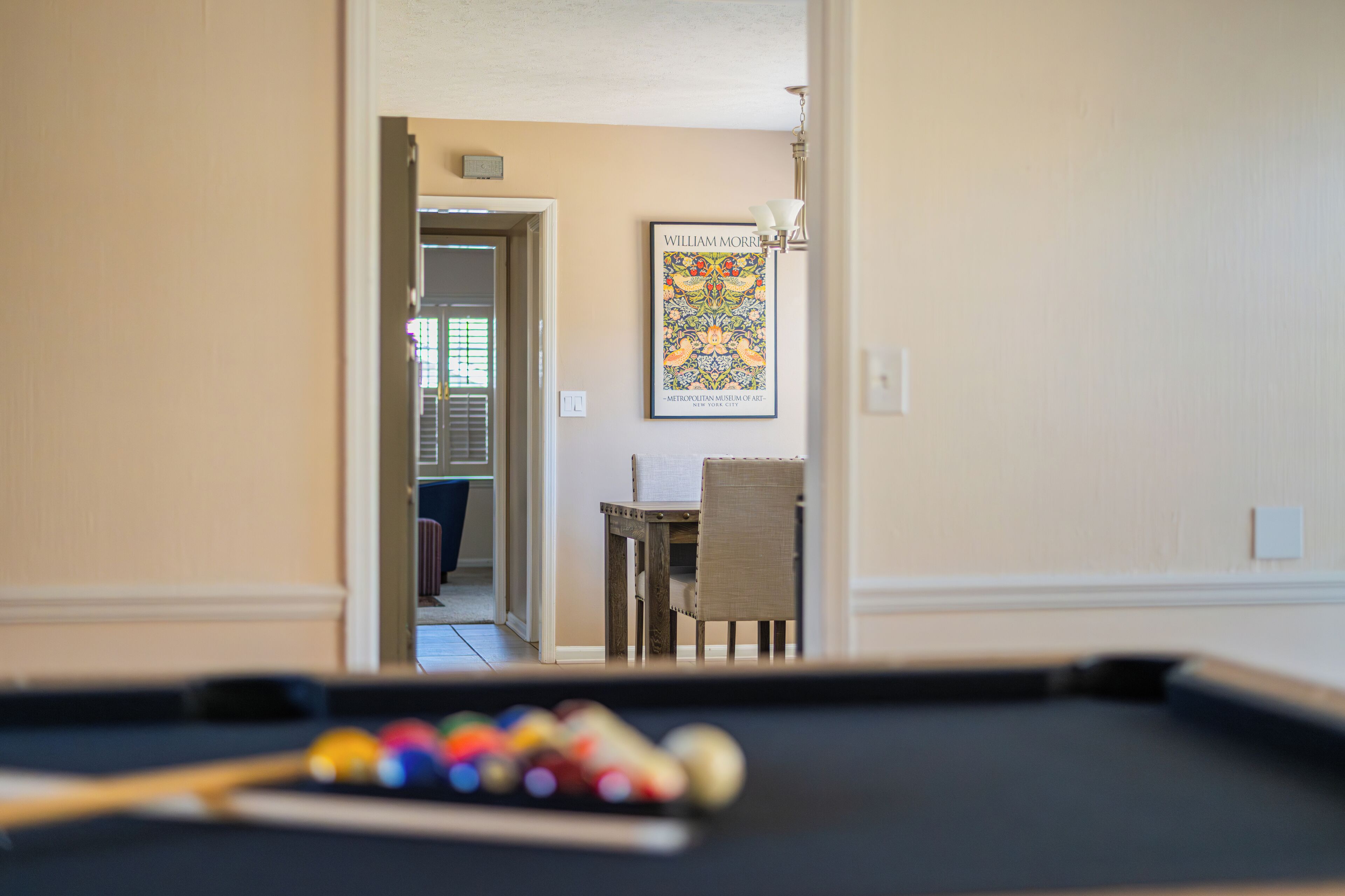 A pool table with balls in the foreground, a dining area with a chair and table in the background, and a framed artwork on the wall.