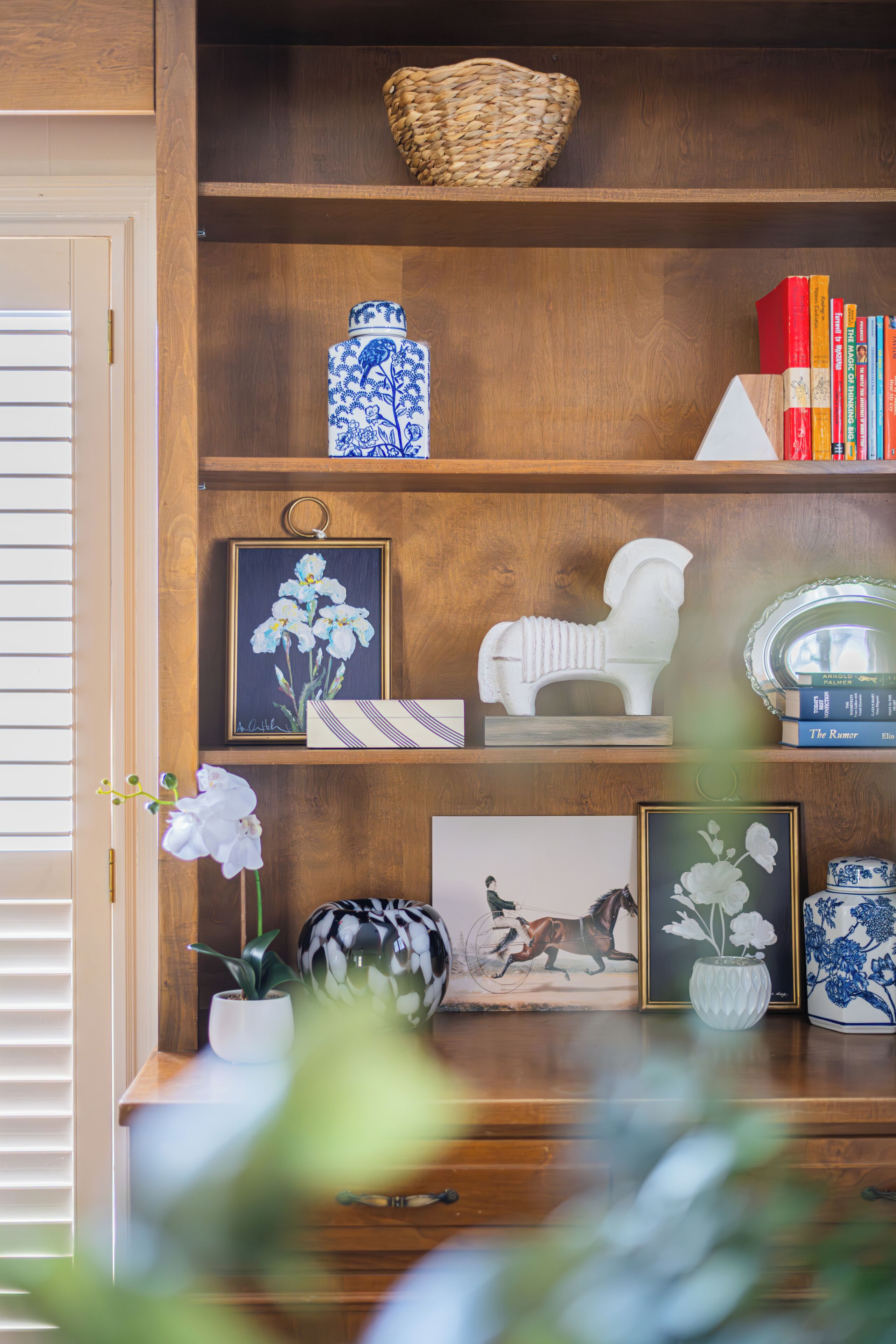 Una estantería de madera con diversos objetos decorativos, incluyendo libros, un florero y obras de arte enmarcadas.