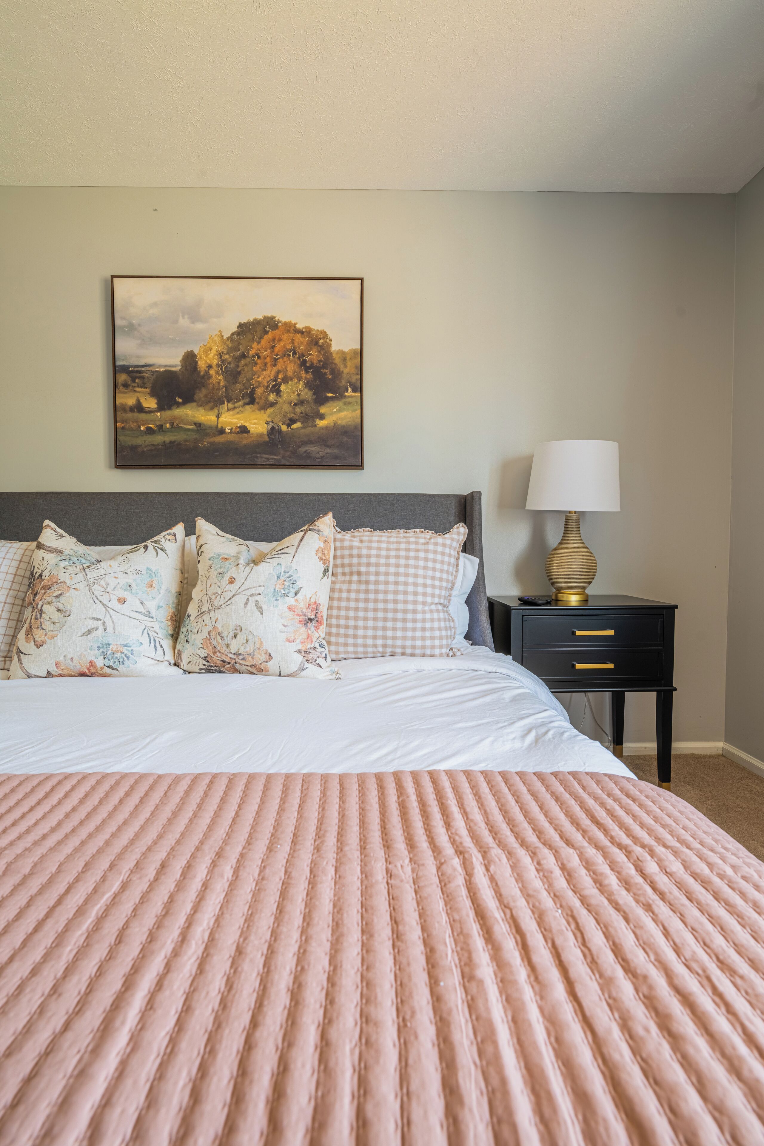 Una cama cuidadosamente tendida con una colcha rosa con textura, un cuadro de paisaje enmarcado encima y una mesita de noche con una lámpara.