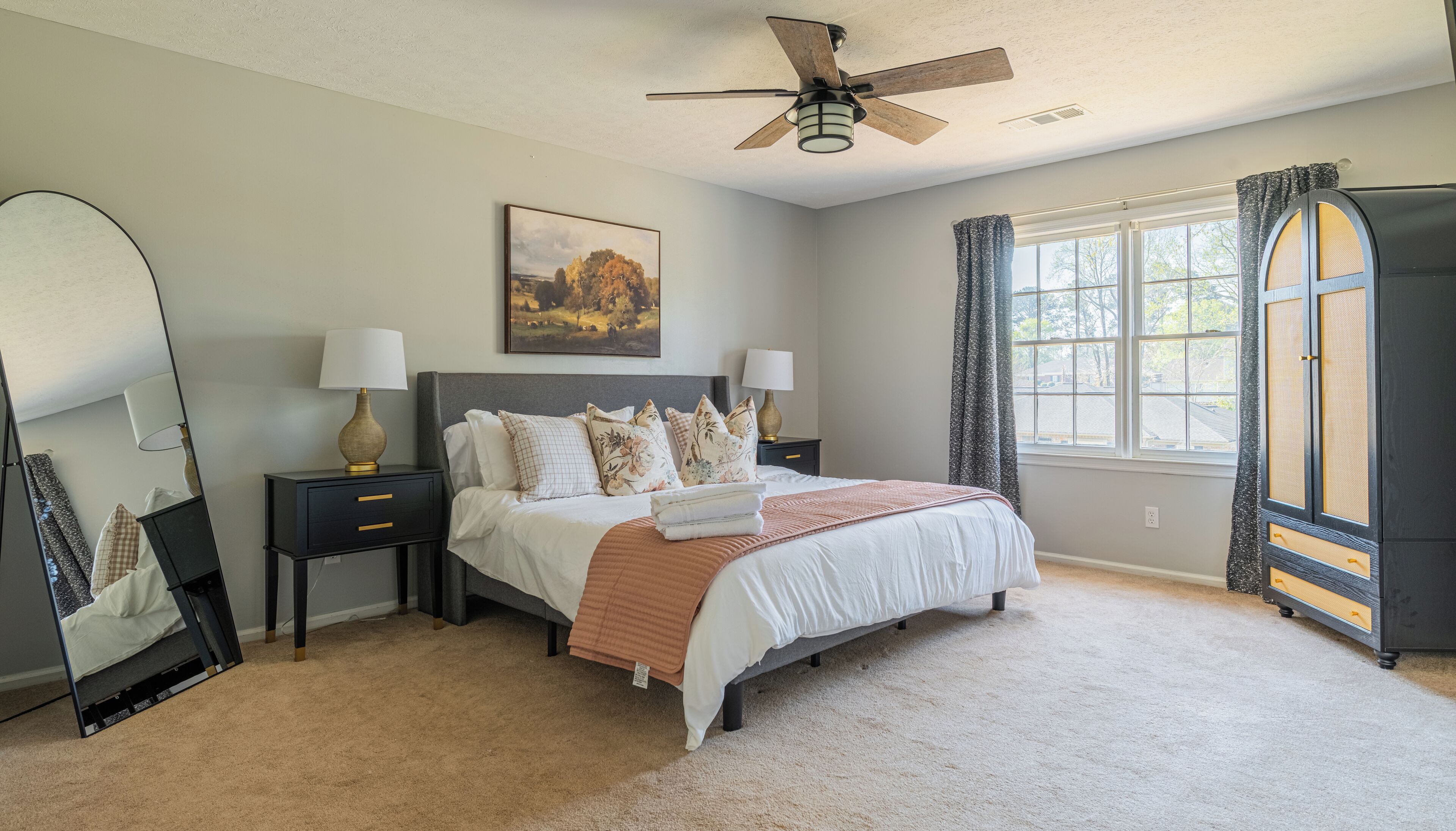 A bedroom with a bed, nightstands, a ceiling fan, a window with curtains, and a large mirror.