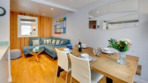 Een moderne woonkamer met een houten vloer, een blauwe bank, een glazen salontafel en een eetgedeelte met een houten tafel en witte stoelen.
