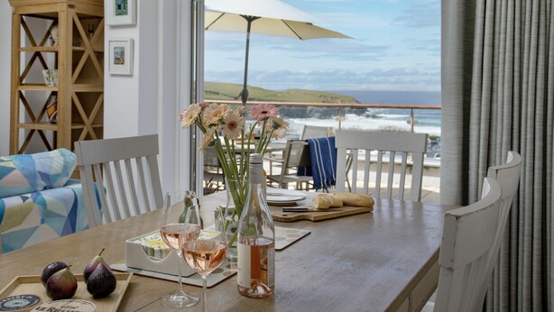 Una mesa de comedor con una botella, copas y un ramillete de flores, con vistas a la costa.
