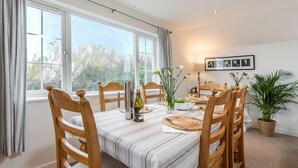 A dining room with a large window, wooden chairs, a striped tablecloth, and a potted plant. - Arnold Cottage is a perfect hideaway for couples, families, or small groups looking to unwind (St. Merryn, Treyarnon Bay)
