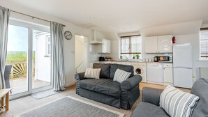 A modern living room with a grey sofa, a dining table, and a kitchen area with white appliances. - Arnold Cottage is a perfect hideaway for couples, families, or small groups looking to unwind (St. Merryn, Treyarnon Bay)