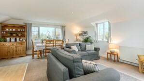 A spacious living room with a grey sofa set, wooden dining table, and a kitchen area with wooden cabinets and a built-in shelf. - Arnold Cottage is a perfect hideaway for couples, families, or small groups looking to unwind (St. Merryn, Treyarnon Bay)