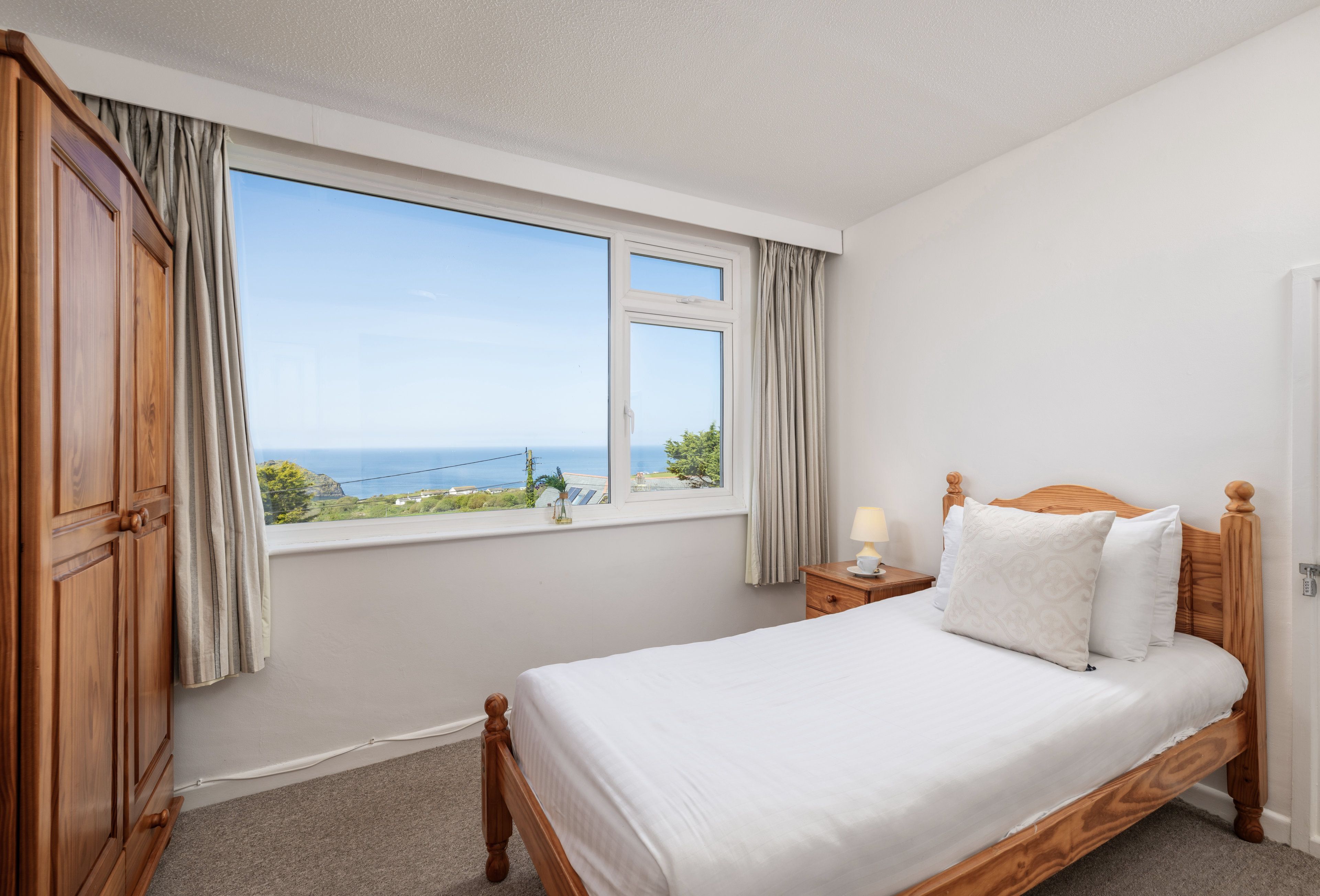 A bedroom with a large window, a wooden bed, a bedside table with a lamp, and a view of the sea.