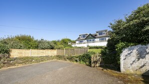 A residential property with a paved driveway, stone wall, wooden fence, and a large house with multiple windows and a sloped roof.