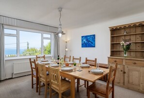 A dining room with a wooden table set for six, surrounded by chairs, with a view of the outdoors through large windows.