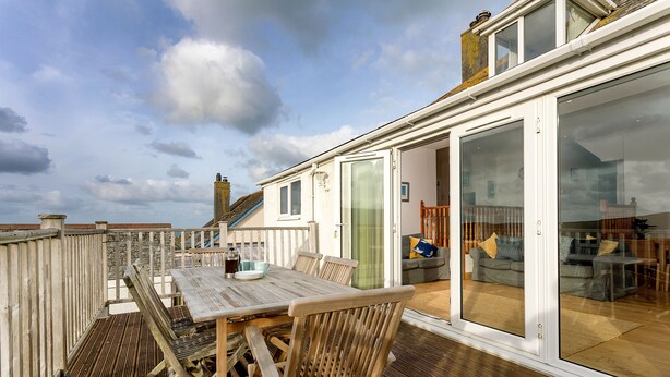 A wooden deck with a table and chairs, a sliding glass door leading to a living area with a sofa and cushions. - Welcome to Bay View, a stunning five-bedroom dog-friendly holiday home (Port Isaac)