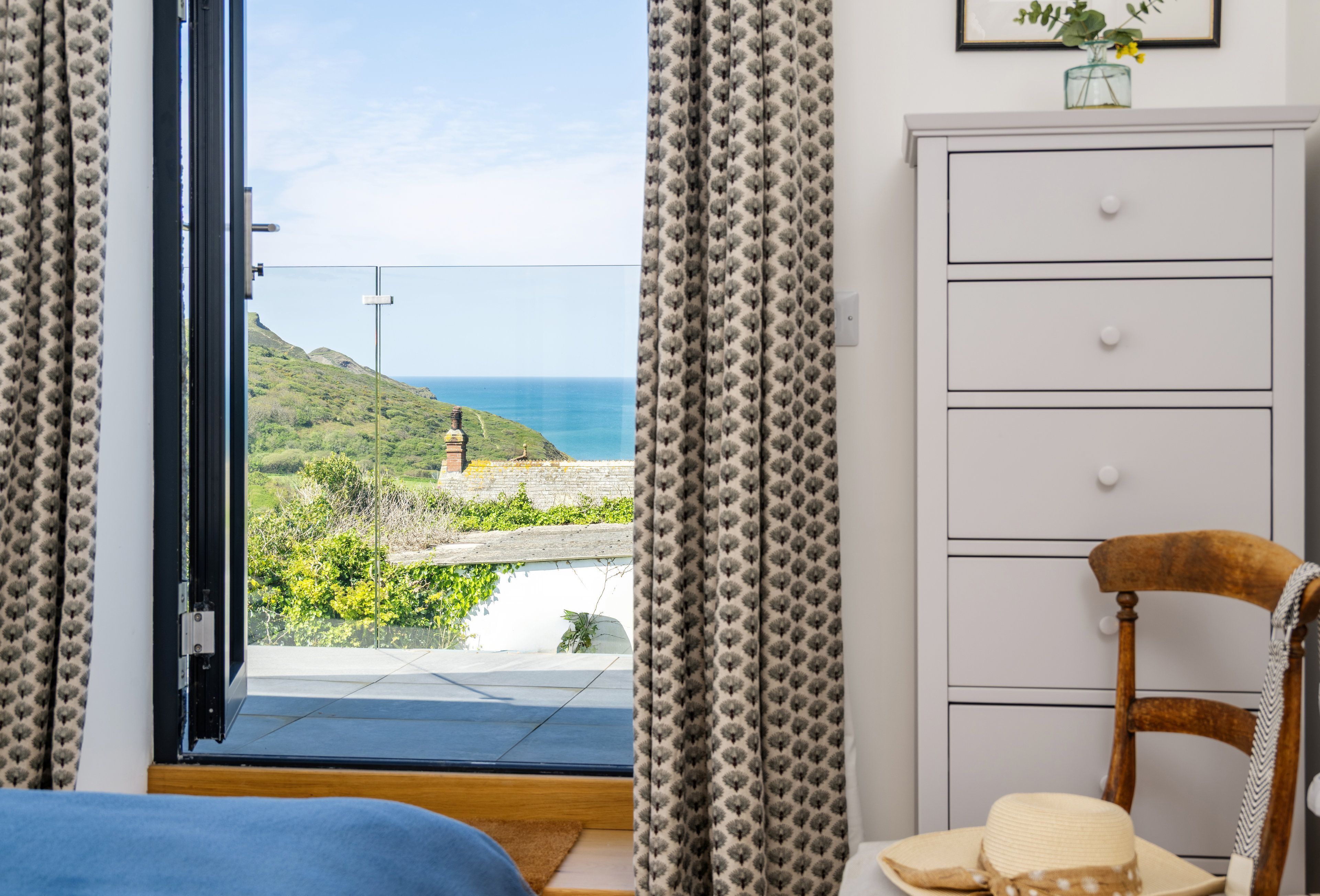 Une chambre avec vue sur la mer, une commode blanche, une chaise en bois et un lit recouvert d’une couverture bleue.