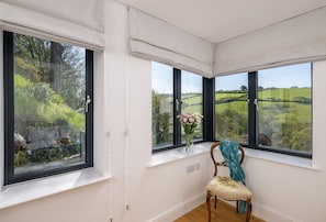 A room with a large bay window overlooking a green landscape, a chair with a floral cushion, and a vase with flowers on the windowsill.