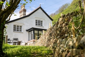 A white house with a stone wall and a chimney. There are two windows and a door.